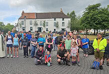 Running group gathering outdoors in a town square, diverse participants with backpacks and sportswear, some with dogs, preparing for a run or walk, under a partly cloudy sky.