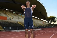 Running athlete smiling on a track at an outdoor stadium, wearing sports gear, ready for training or competition, with a clear blue sky and stadium seating in the background.