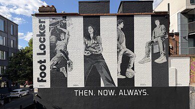Black and white mural of four people sitting and standing, with 'Foot Locker' text and 'Then. Now. Always.' slogan on a city wall.