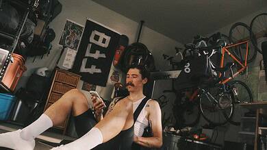 Man with a mustache and white socks sitting on the floor in a garage gym, looking at his smartphone, surrounded by cycling gear and equipment.